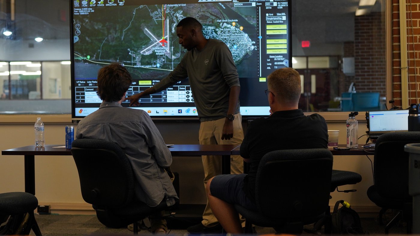 A USI instructor briefing a crew in front of a large operations display showing a map and flight data